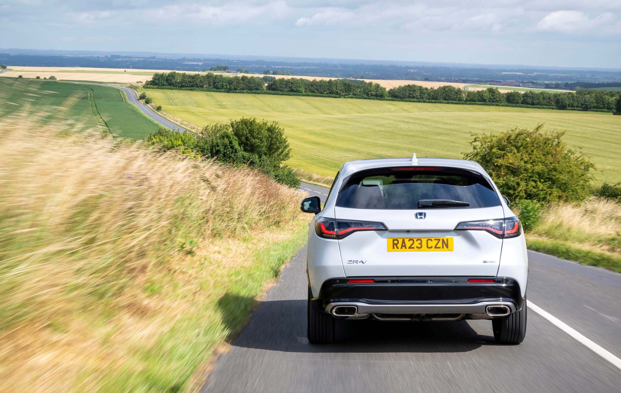 A white Honda ZR-V drives down a rural road flanked by fields and hedges under a partly cloudy sky.