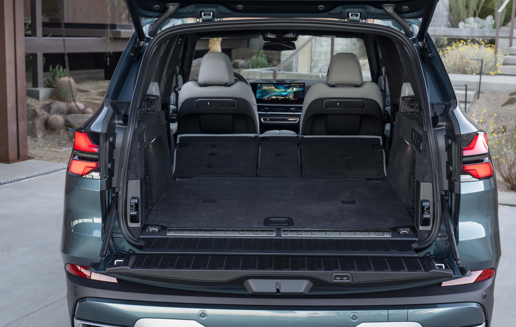 BMW X5 with rear hatch open, showing spacious boot area with folded rear seats, beige interior, and a view of the front dashboard screen.