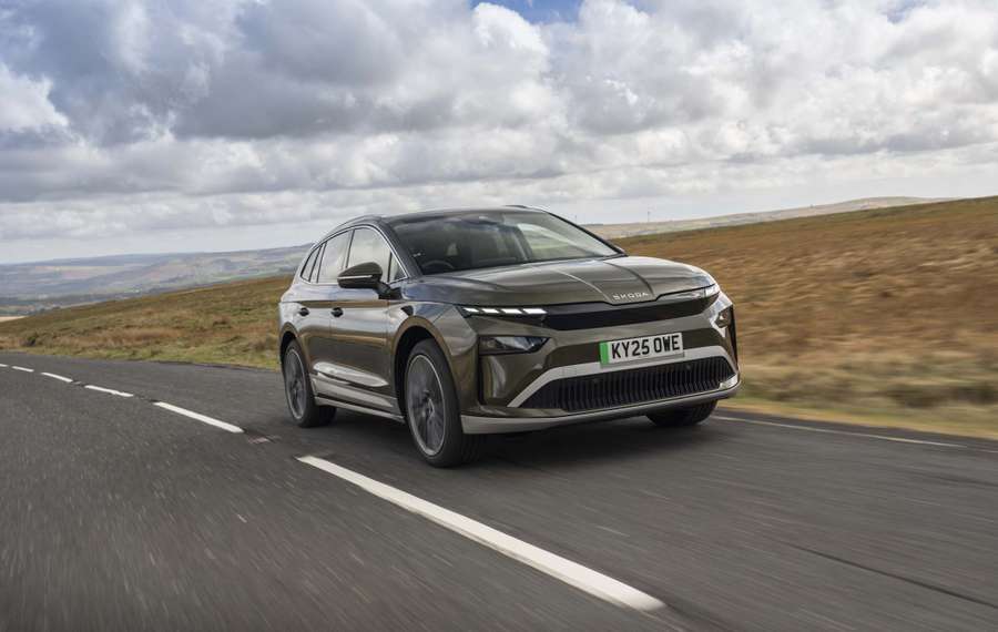 A Škoda Enyaq drives on a rural road under a partly cloudy sky, surrounded by open fields and distant hills.