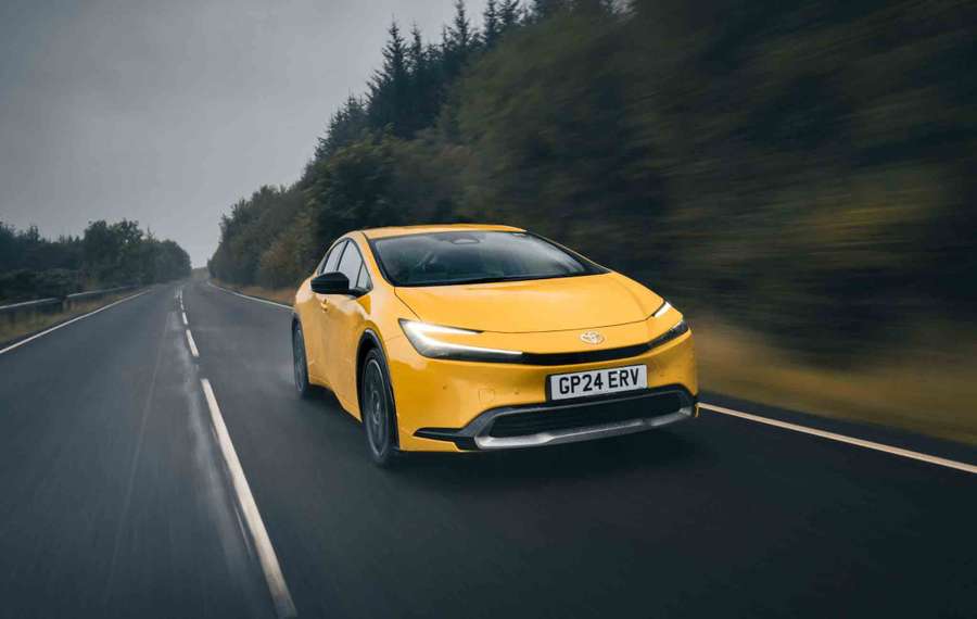 A yellow Toyota Prius drives on a wet, empty road surrounded by blurred greenery, under a cloudy sky.
