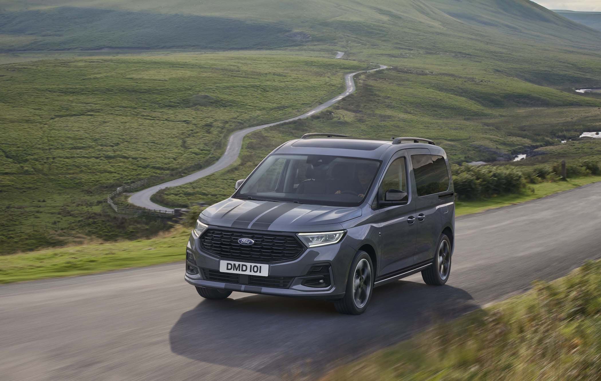 A grey Ford Tourneo Connect drives on a winding road through lush green hills under a clear sky.