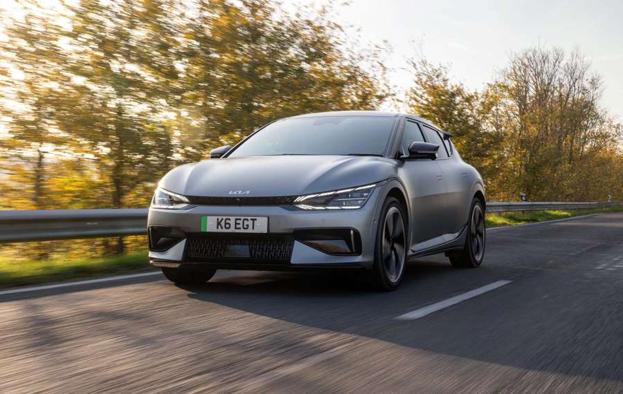 A grey Kia EV6 GT drives on a scenic road with autumn trees in the background under a clear sky.
