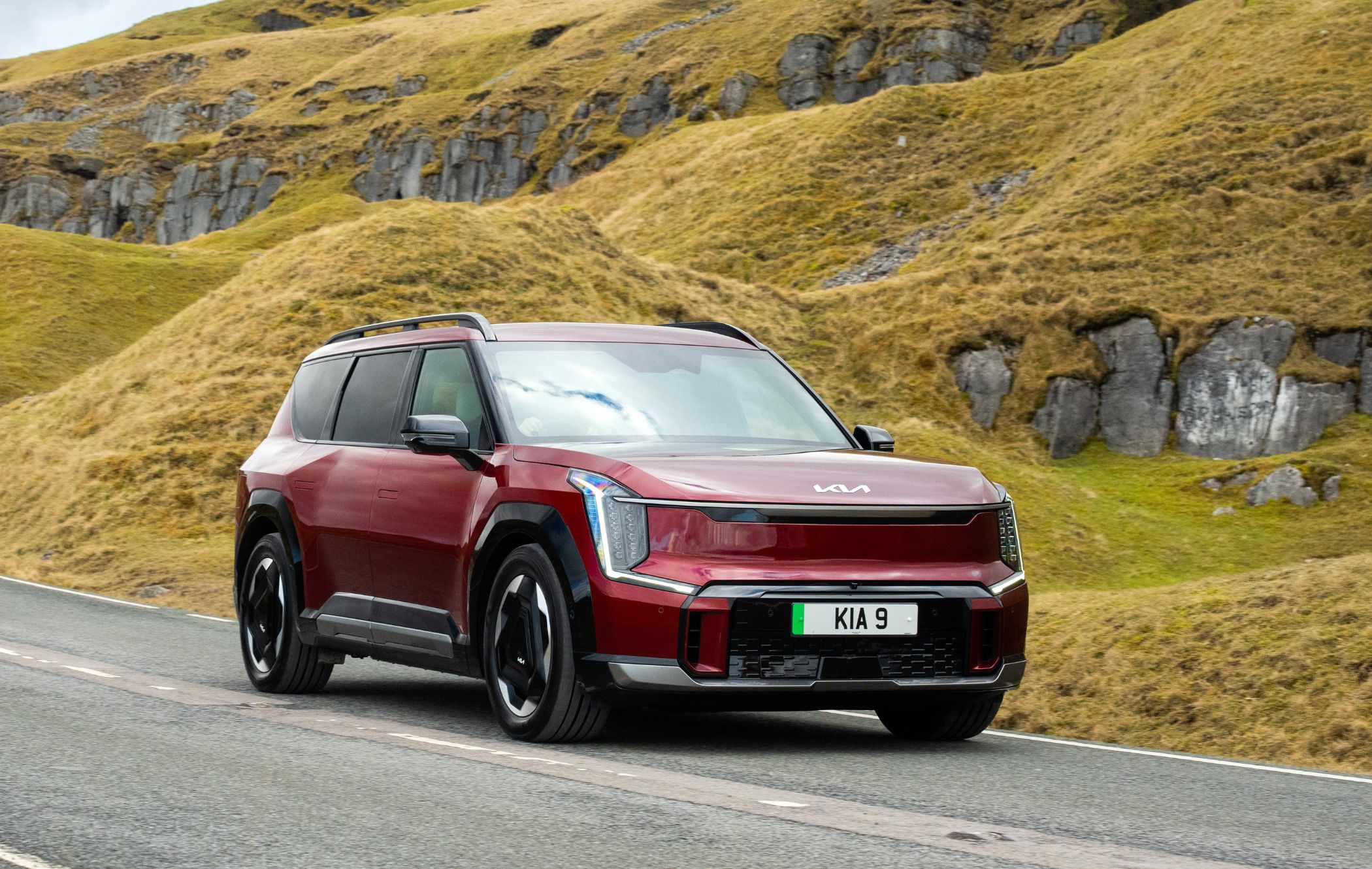 Red Kia SUV driving on a scenic road with grassy hills in the background.