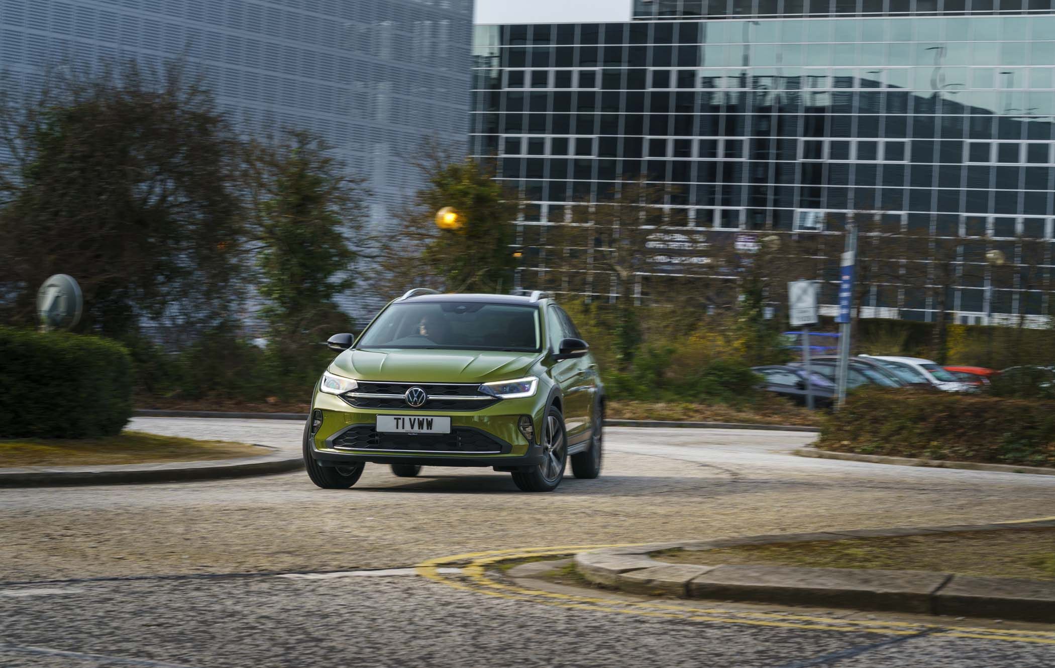 A green Volkswagen Taigo drives on a roundabout in an urban area, with glass buildings and trees in the background.