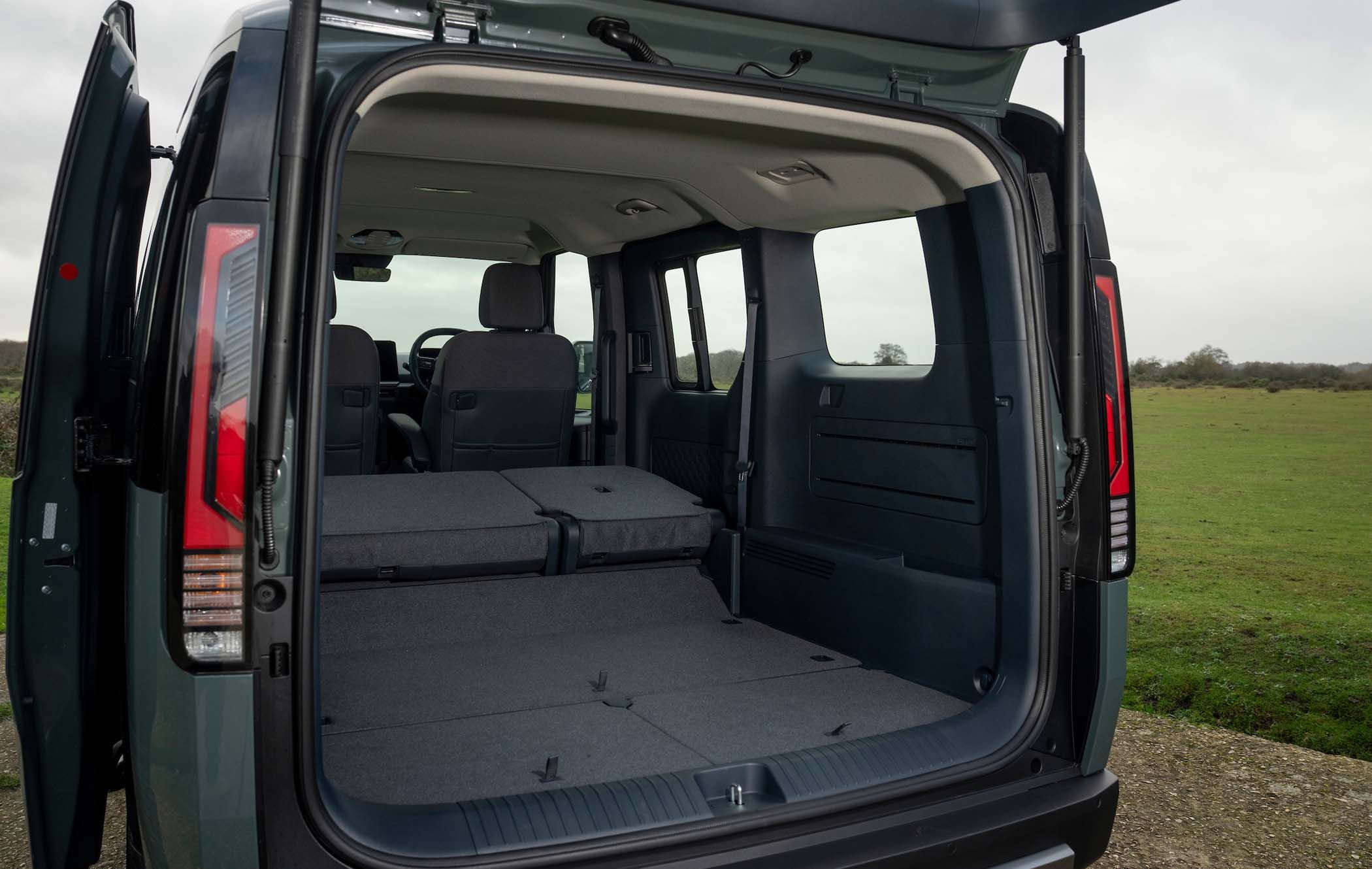 Open rear view of a Kia PV5 Passenger with folded back seats, revealing a spacious luggage area. The scene is outdoors with a grassy landscape in the background.