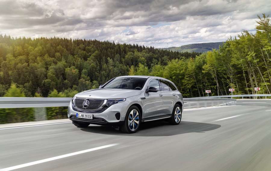 Silver Mercedes EQC driving on a highway through a forested area under a cloudy sky.