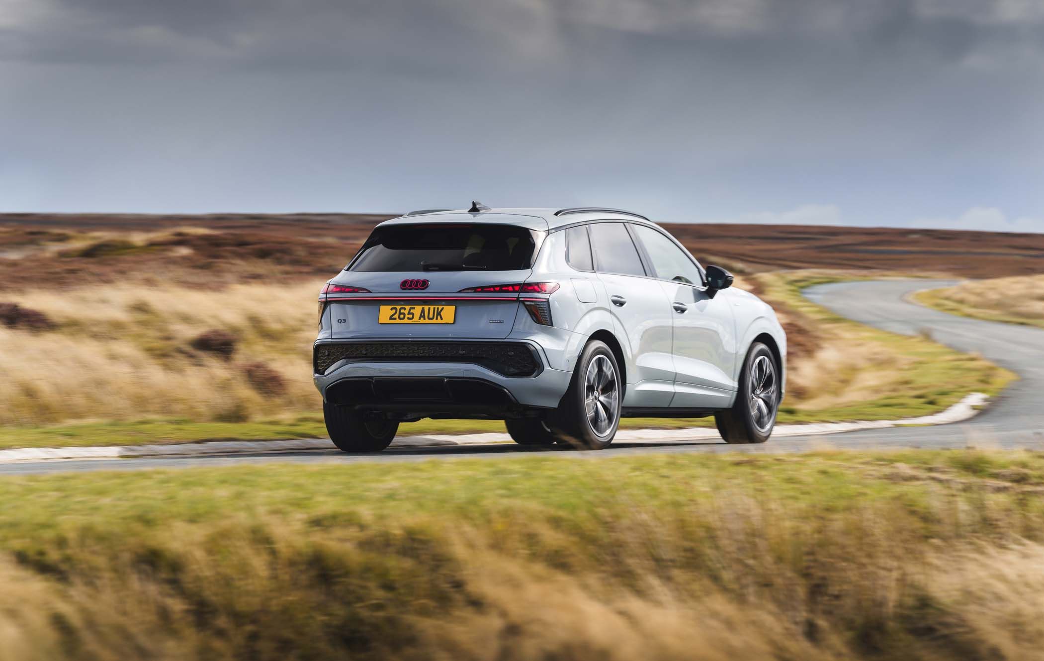 Silver Audi Q3 driving on a winding rural road surrounded by tall grass under a cloudy sky.