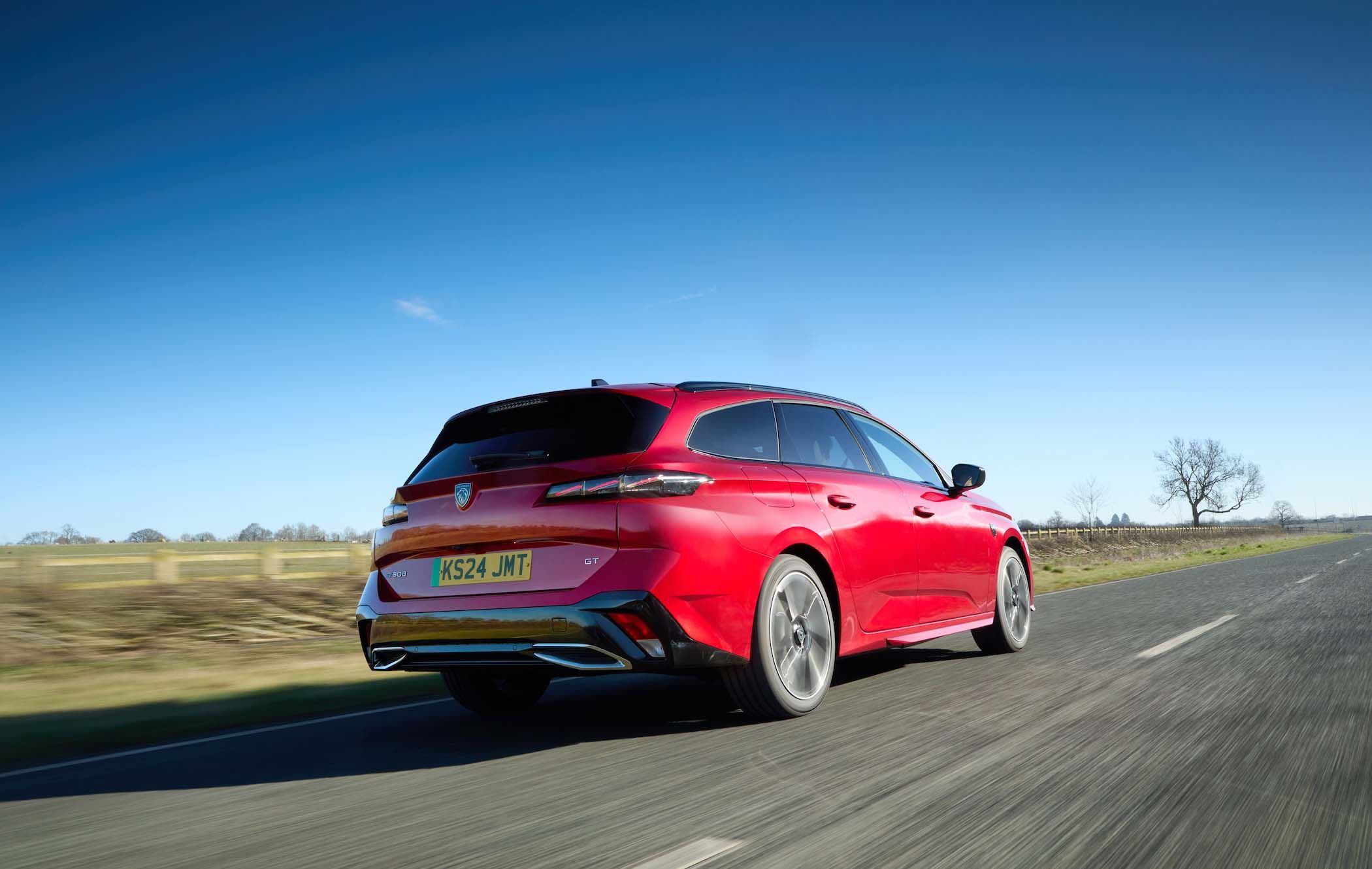 Red Peugeot e-308 SW driving on a rural road under a clear blue sky, with fields and trees in the background.