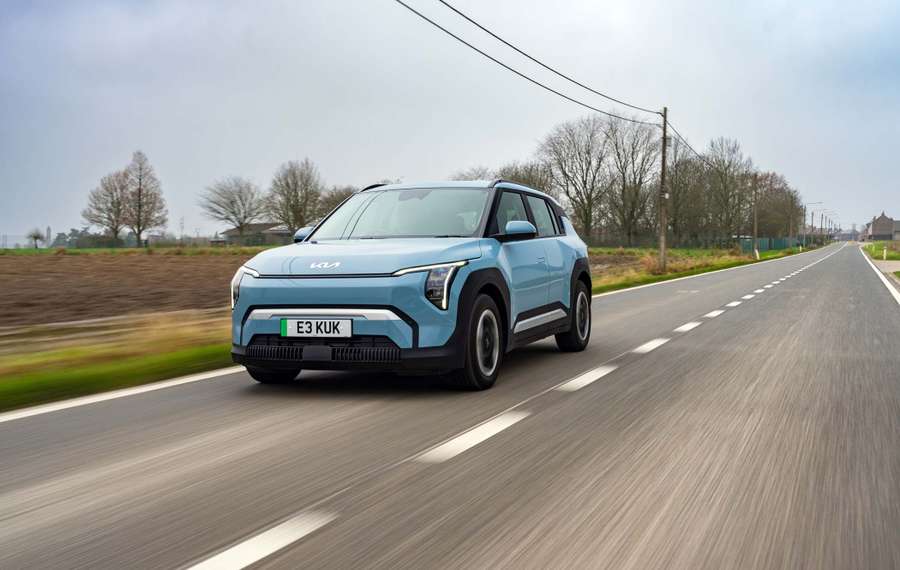 A light blue Kia EV3 drives on a rural road, surrounded by fields and leafless trees under a cloudy sky.