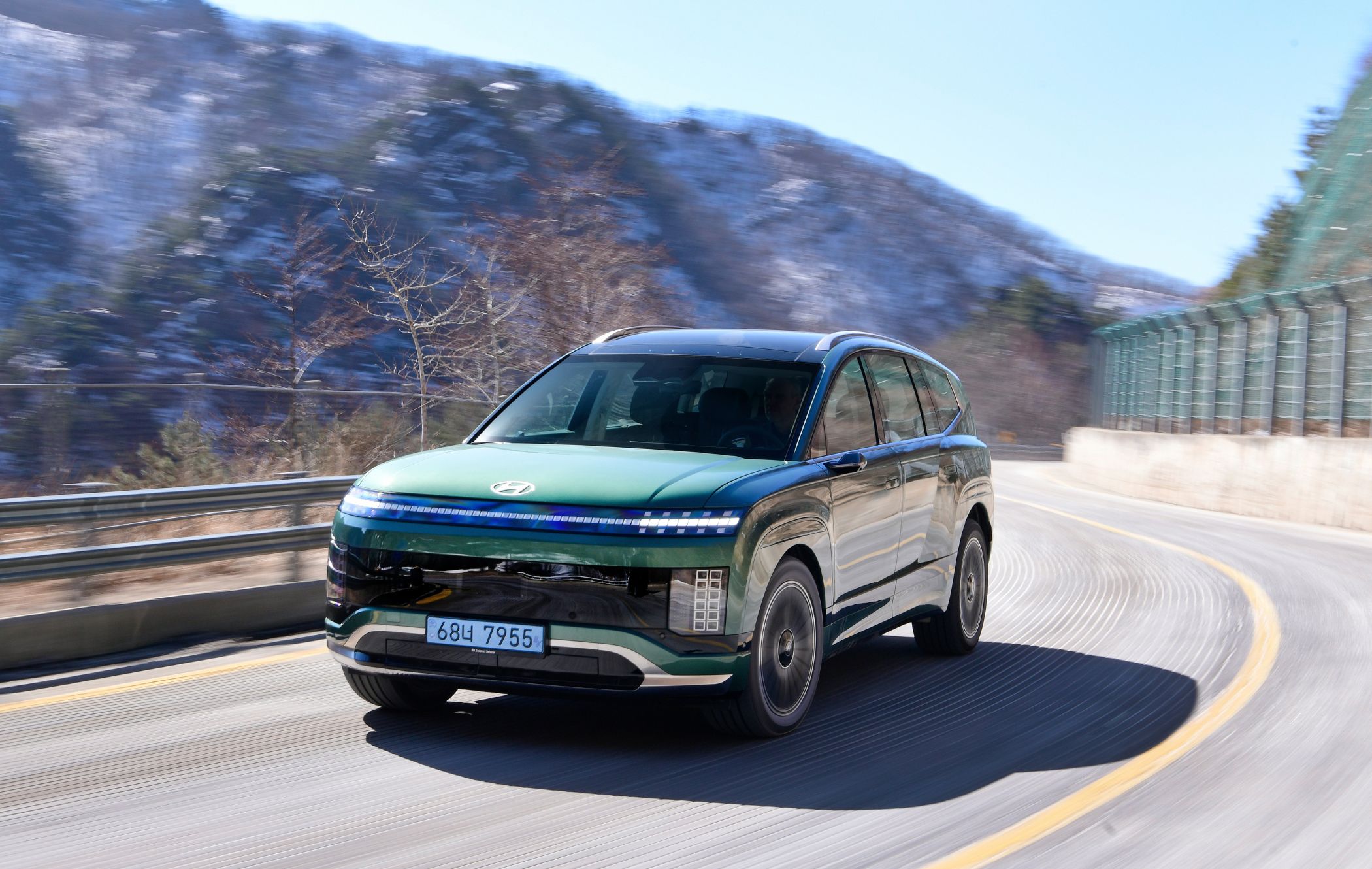 A green Hyundai IONIQ 9 drives on a winding mountain road with snow-covered hills in the background under a clear blue sky.