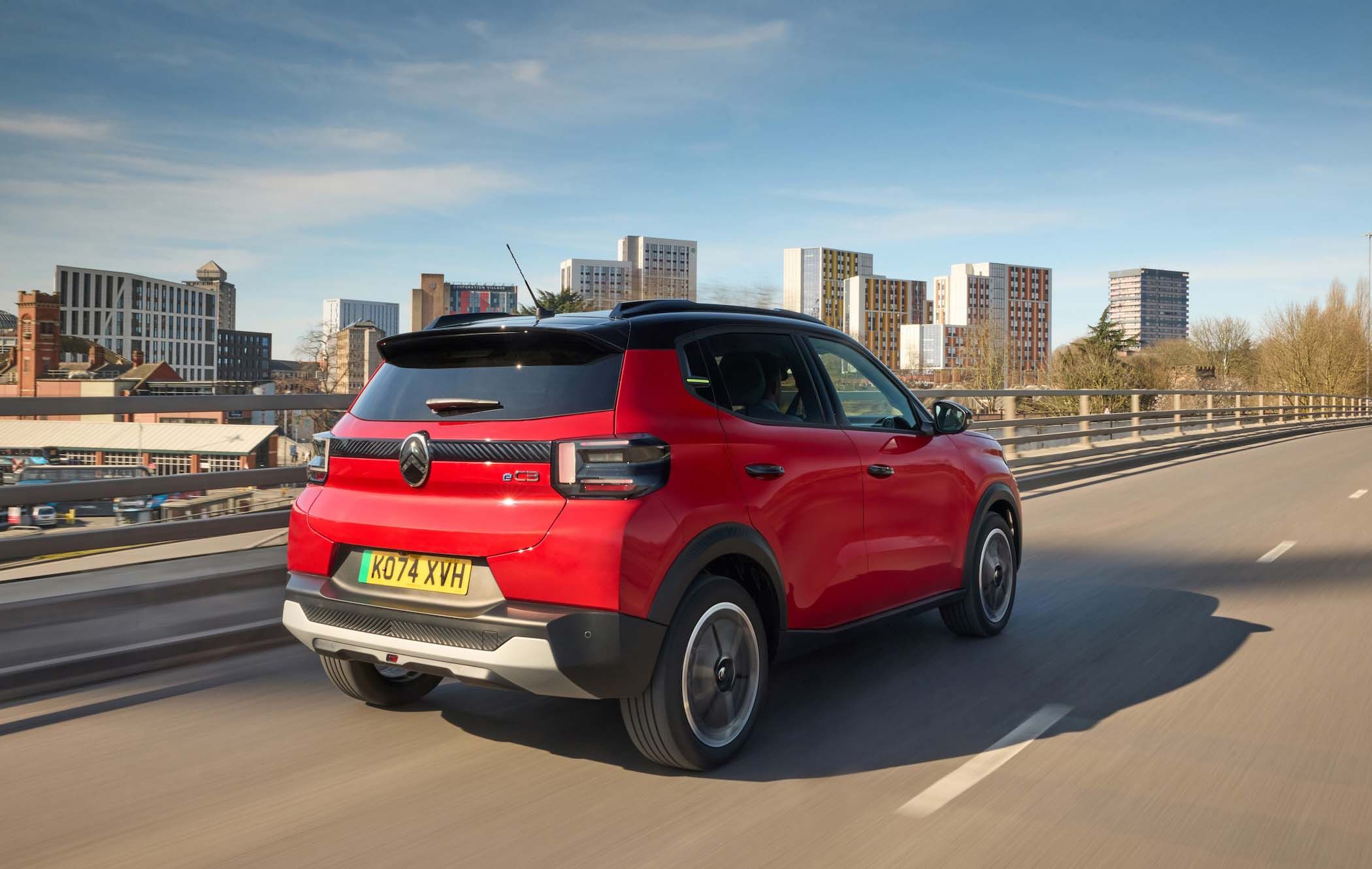 Red Citroën ë-C3 driving on a highway with city buildings in the background under a clear blue sky.