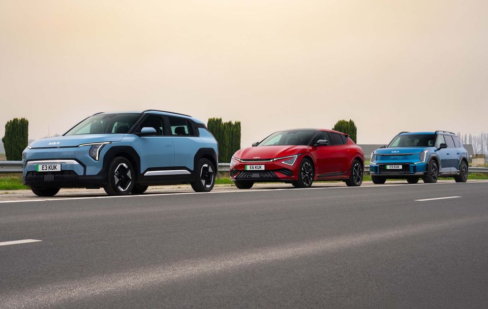 One blue Kia EV3, one red Kia EV6, and a blue Kia EV9 parked in a row on a road with trees in the background.