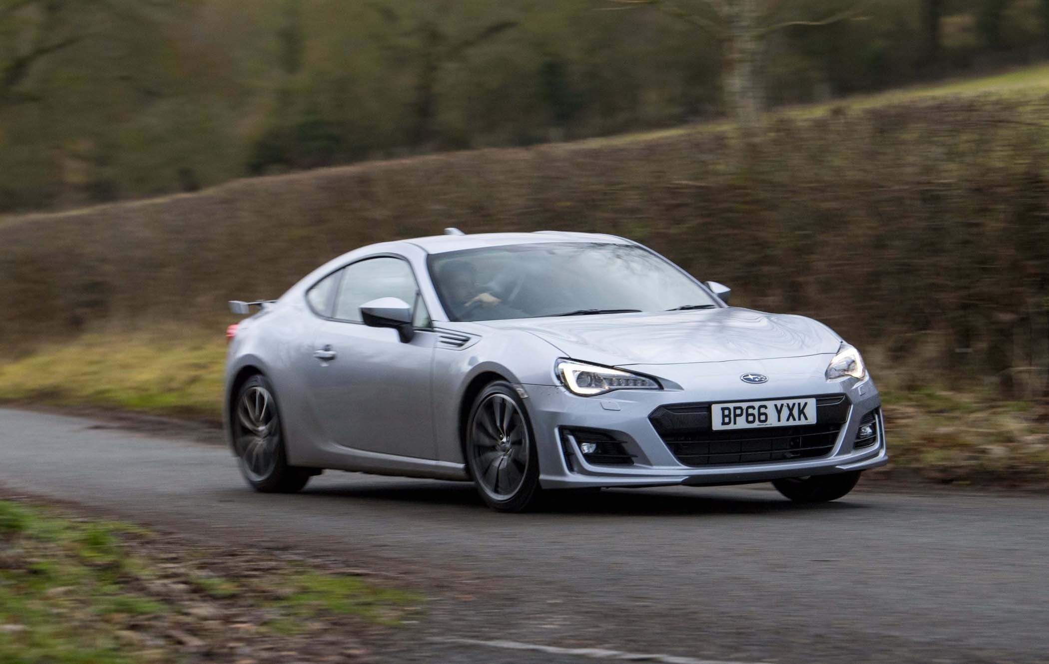 Silver Subaru BRZ driving on a rural road, surrounded by hedges and trees, with a person visible inside.