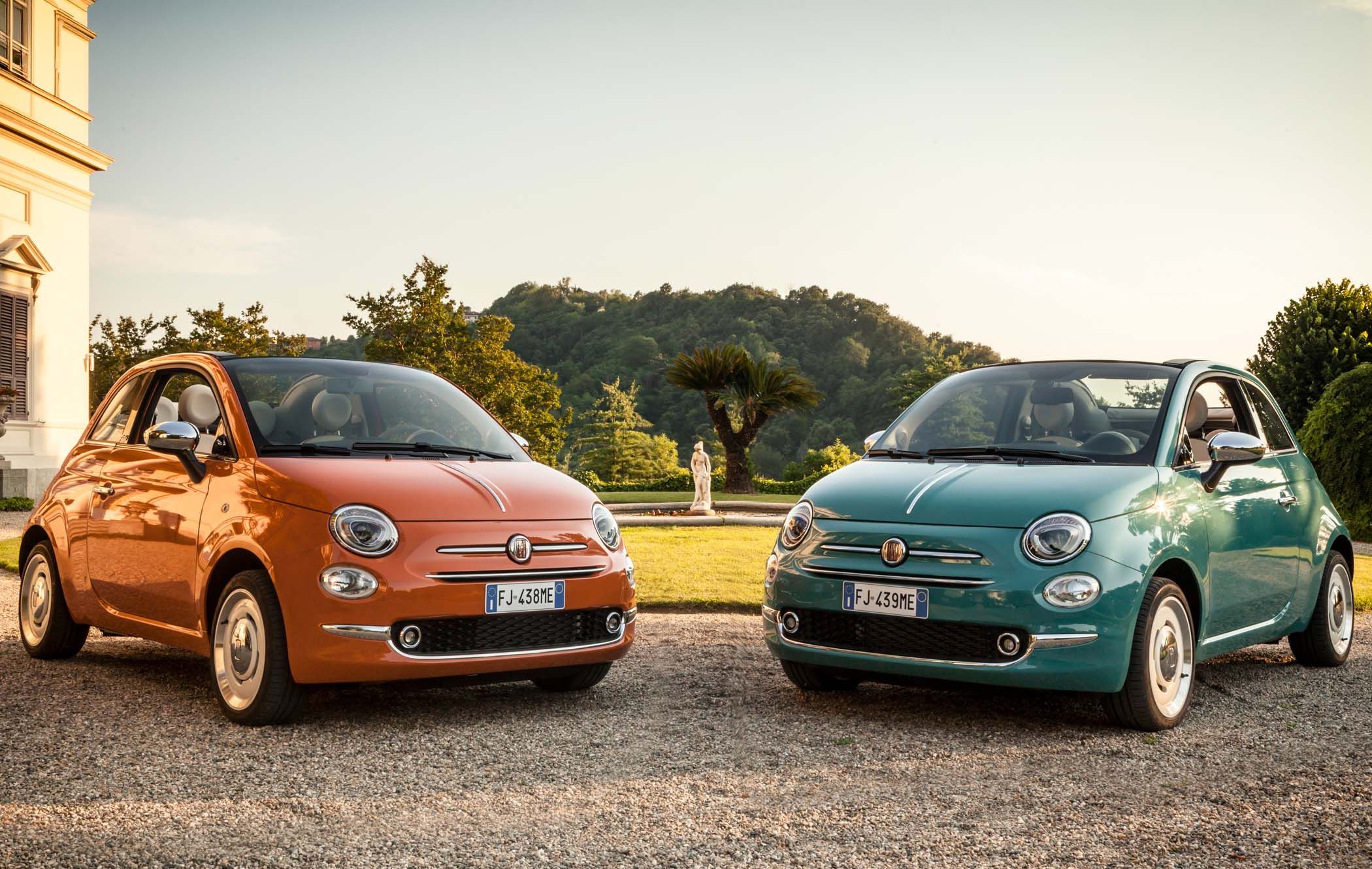 Two Fiat 500 cars, one orange and one teal, parked on a gravel path in front of greenery and a statue, with a clear sky overhead.
