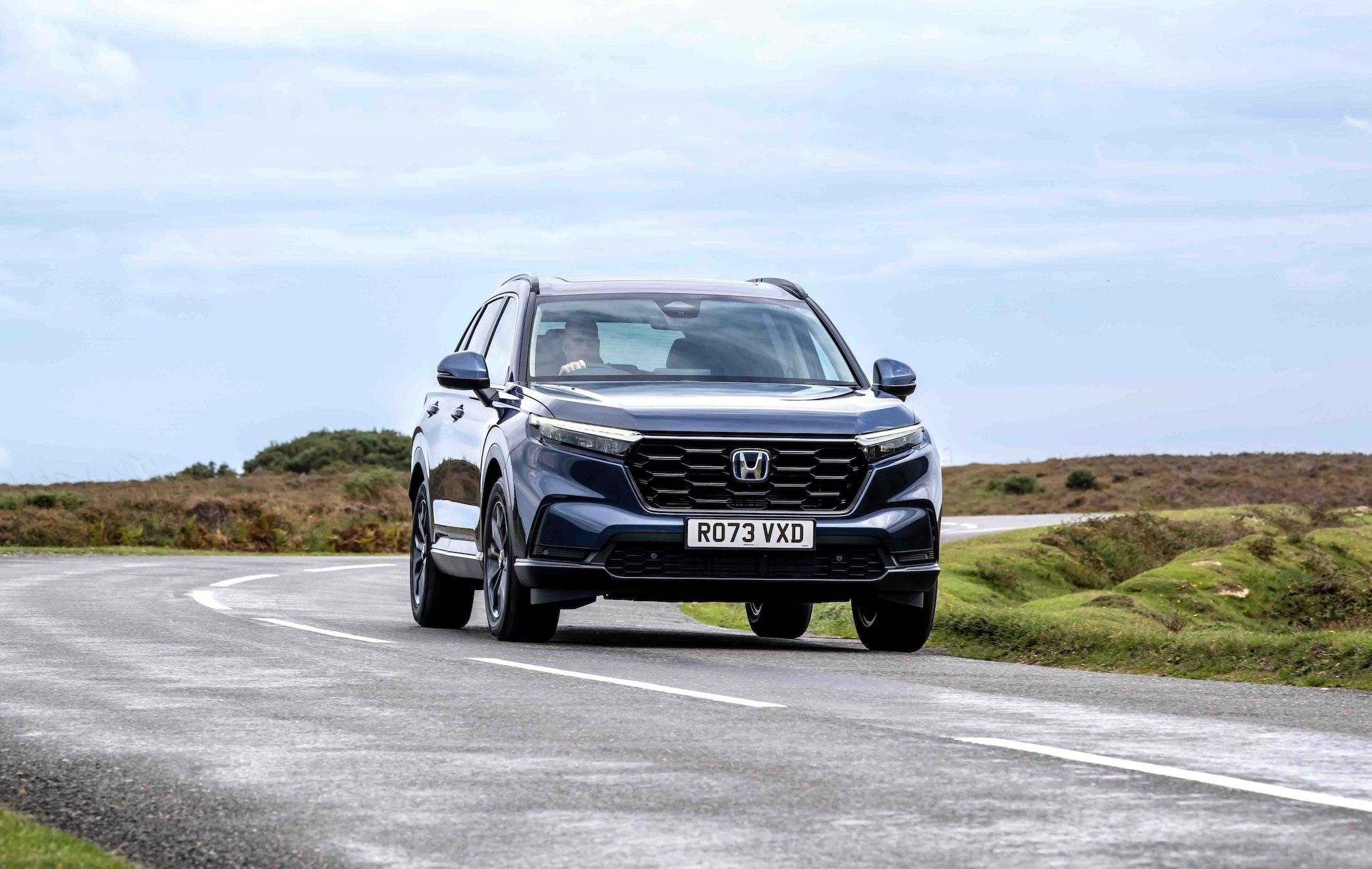A blue Honda CR-V drives on a winding road through a scenic landscape with cloudy skies.