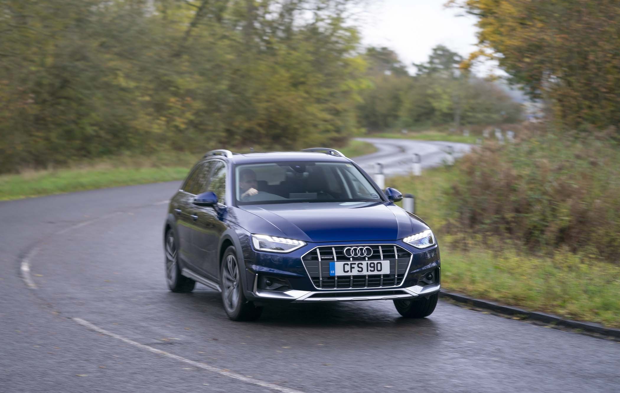 A blue Audi A4 Allroad driving on a winding road surrounded by greenery and autumn trees.