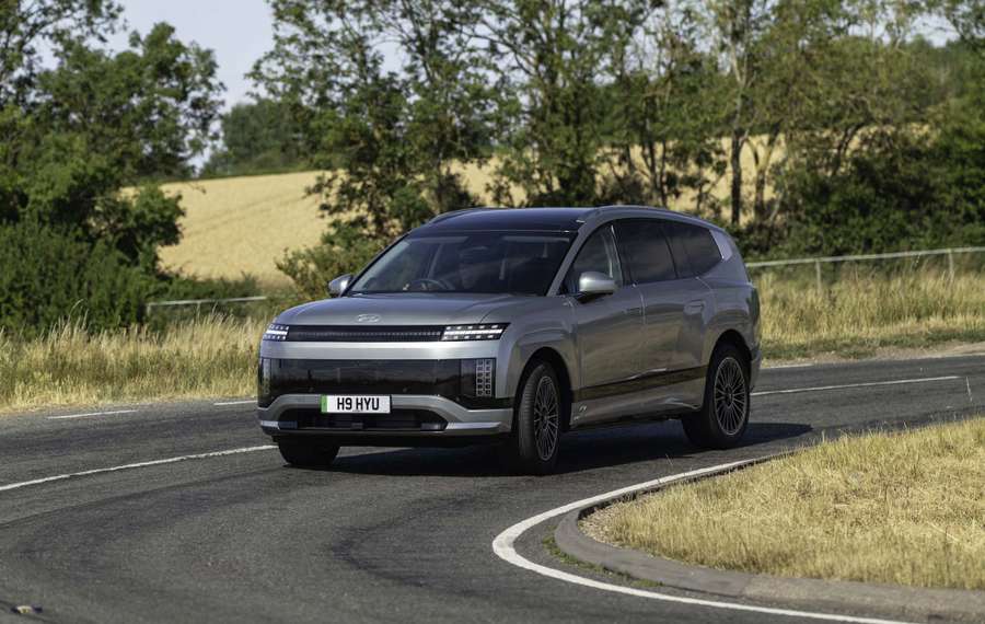 Silver Hyundai IONIQ 9 driving on a curved rural road, surrounded by trees and fields under a clear sky.