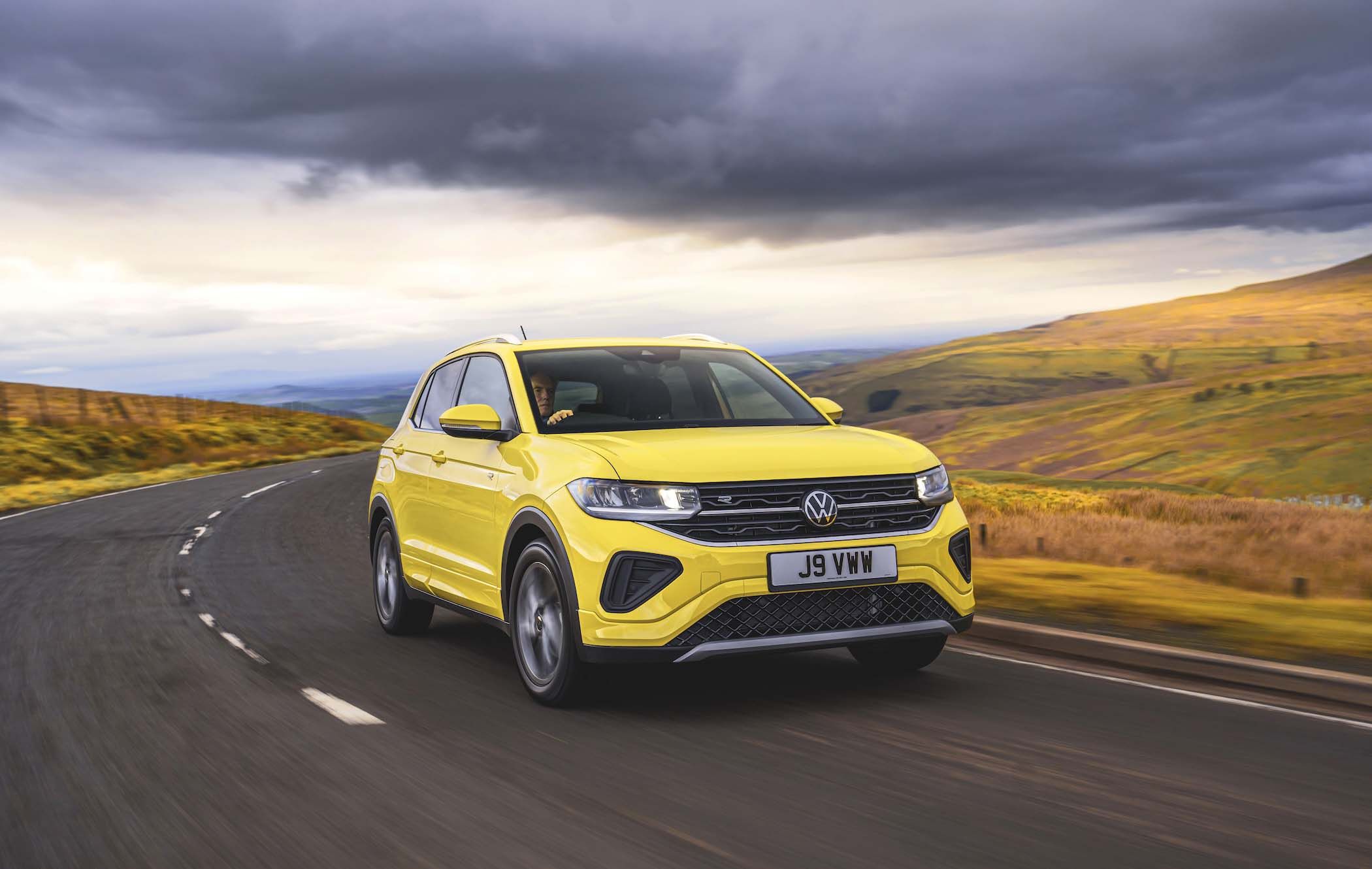 A bright yellow VW T-Cross drives on a winding road through a scenic landscape with rolling hills and a cloudy sky.