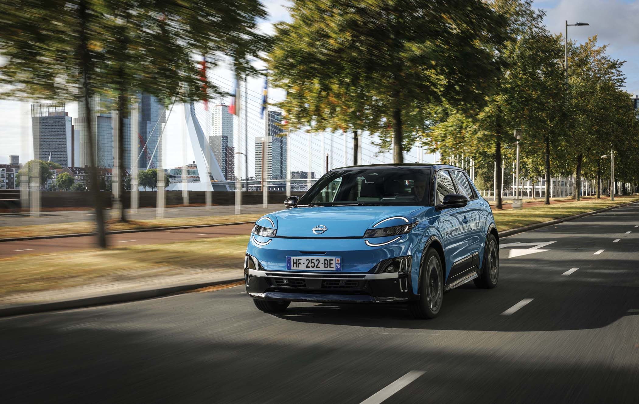 Blue Nissan MICRA driving on a city road with a modern bridge and skyscrapers in the background, surrounded by trees.