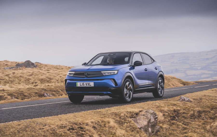 A blue Vauxhall Mokka Electric drives on a rural road through a hilly landscape under a cloudy sky.