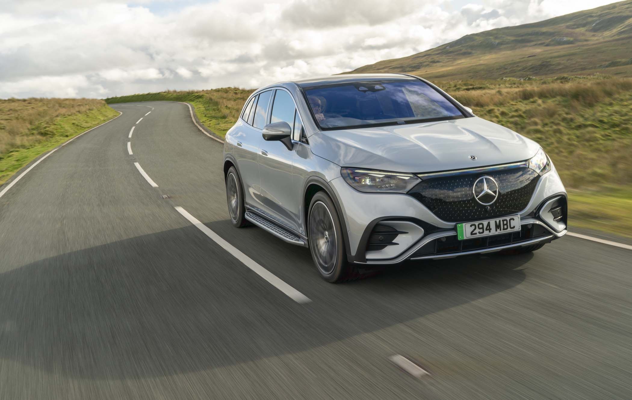 Silver Mercedes EQE SUV driving on a winding road through a scenic countryside with hills and a cloudy sky.