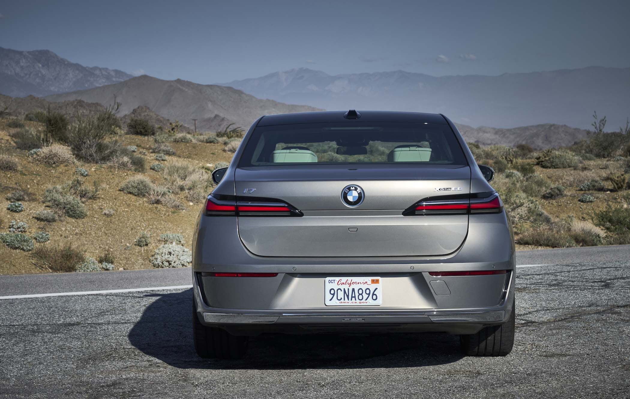 Rear view of a silver BMW 7 Series parked on a desert road, with mountains in the background under a clear sky.