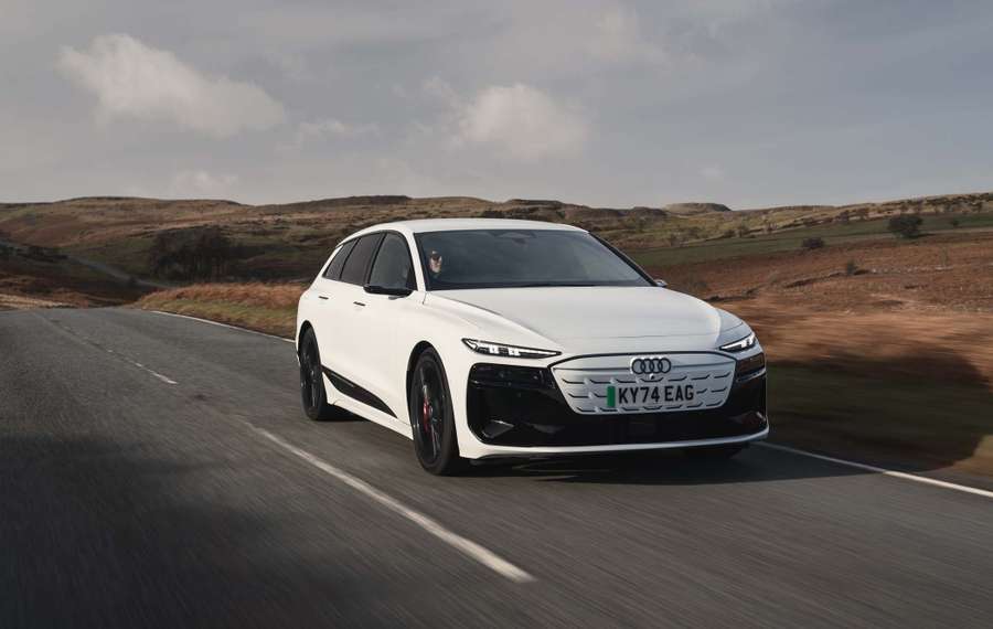 White Audi S6 Avant e-tron driving on a rural road with hills and a cloudy sky in the background.