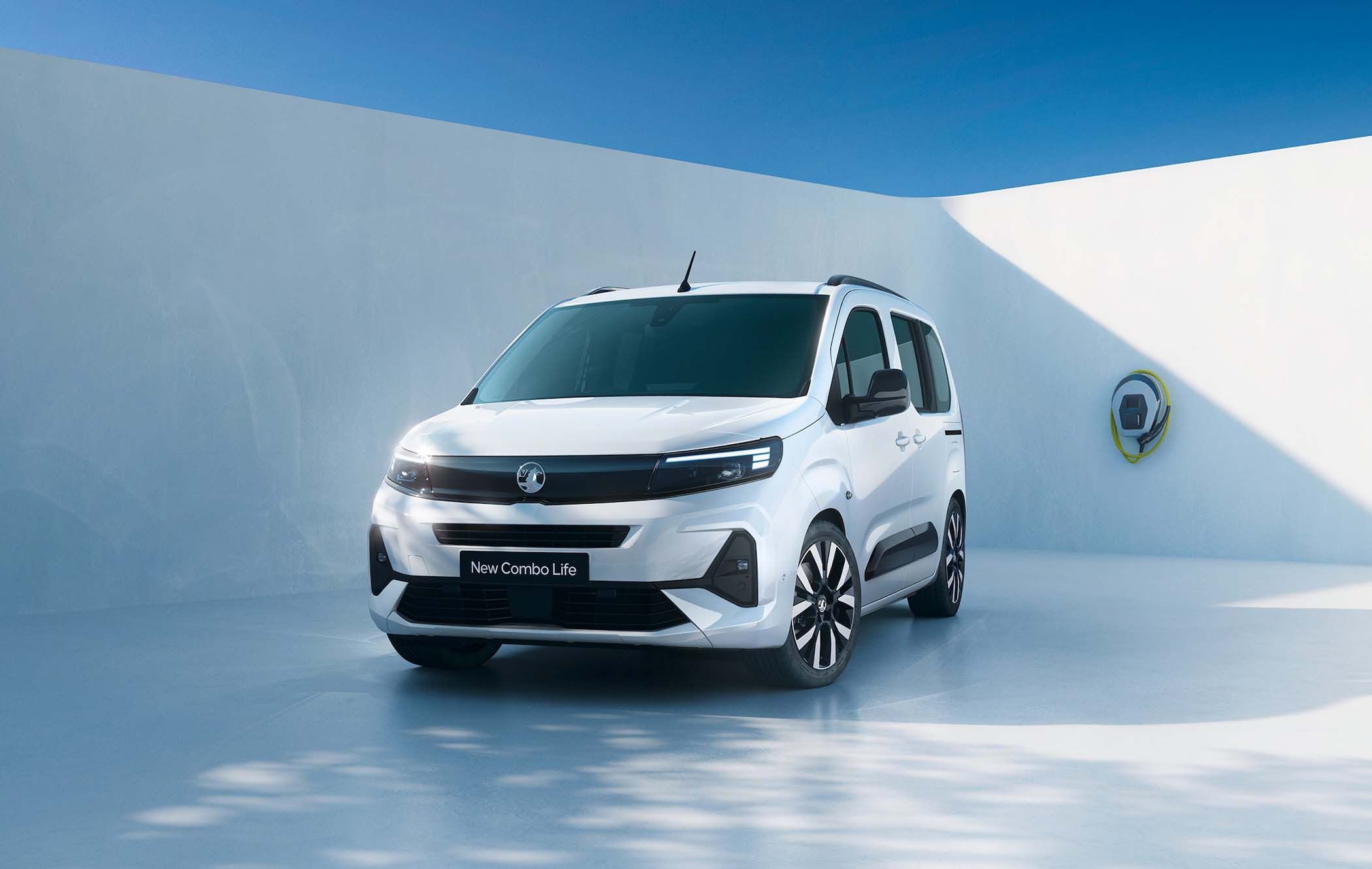 White Vauxhall Combo Life parked in a modern, sunlit courtyard with a minimalist wall and an electric vehicle charging point.