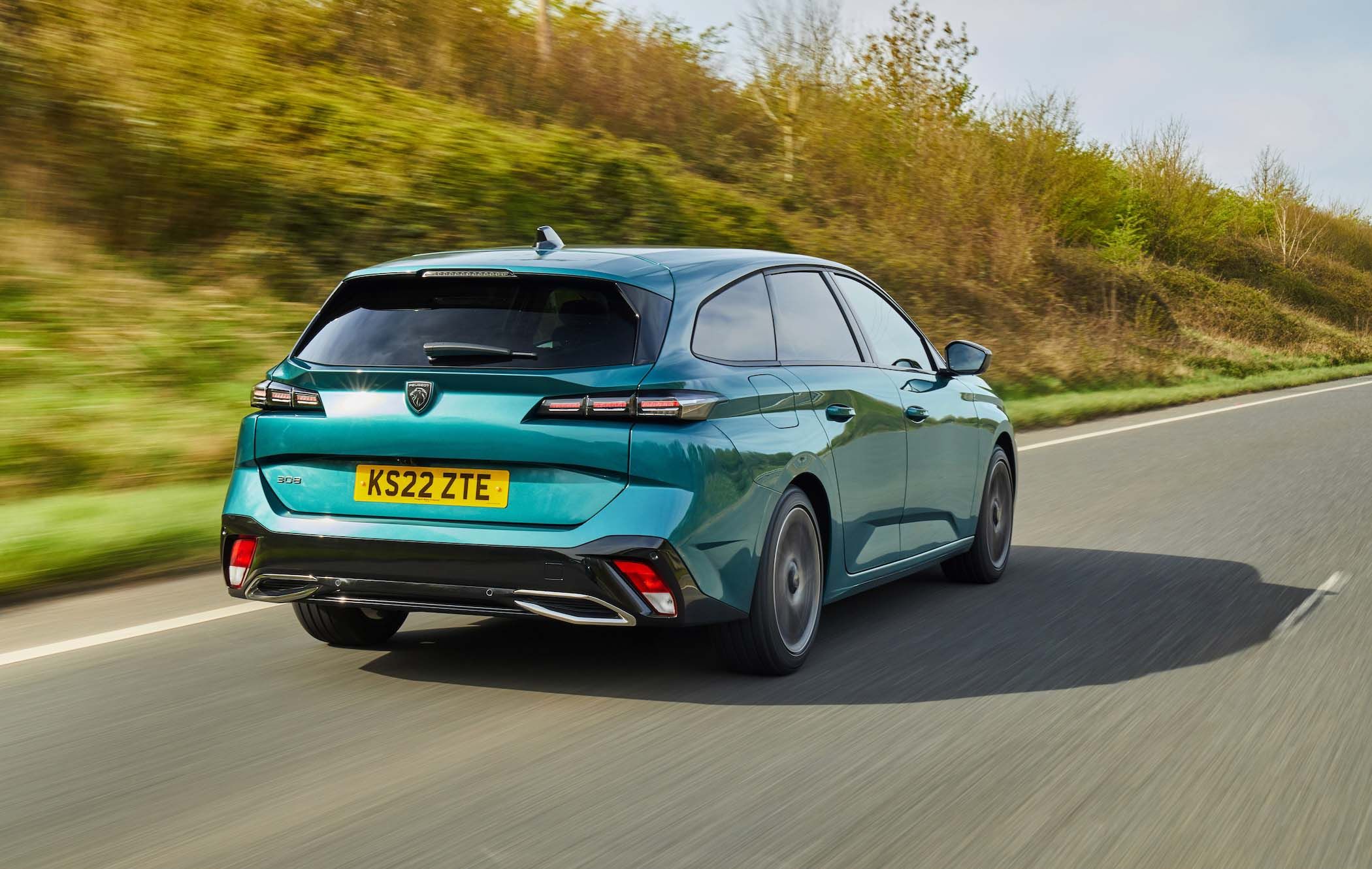 A teal Peugeot 308 SW driving on a rural road, surrounded by greenery and a clear sky.