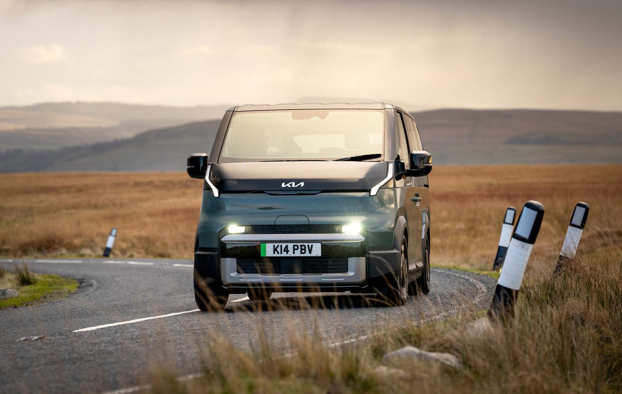 A Kia PV5 Passenger drives on a winding rural road, surrounded by open fields under a cloudy sky.