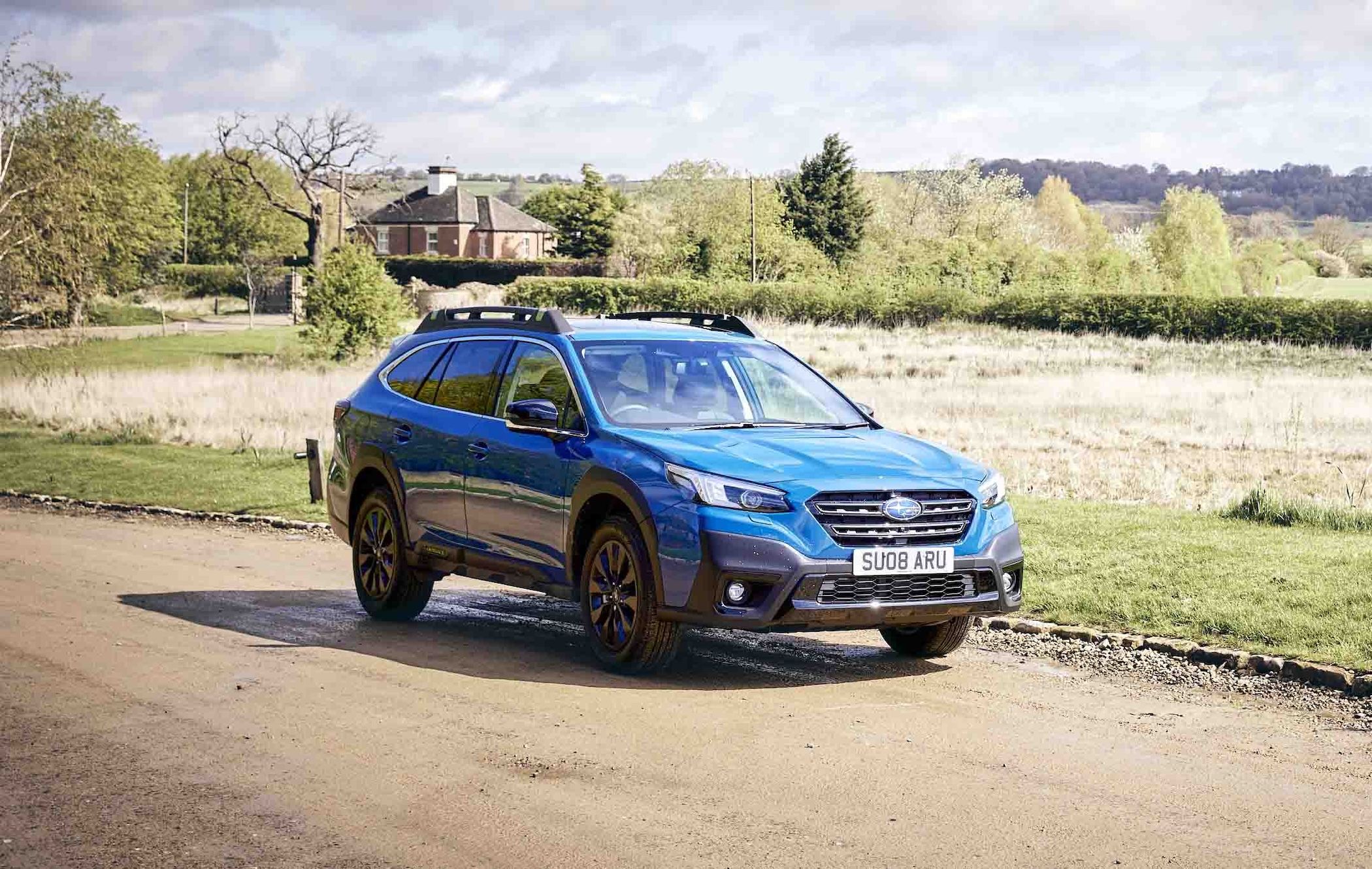 A blue Subaru Outback parked on a rural dirt road, with a house and lush green landscape in the background under a partly cloudy sky.