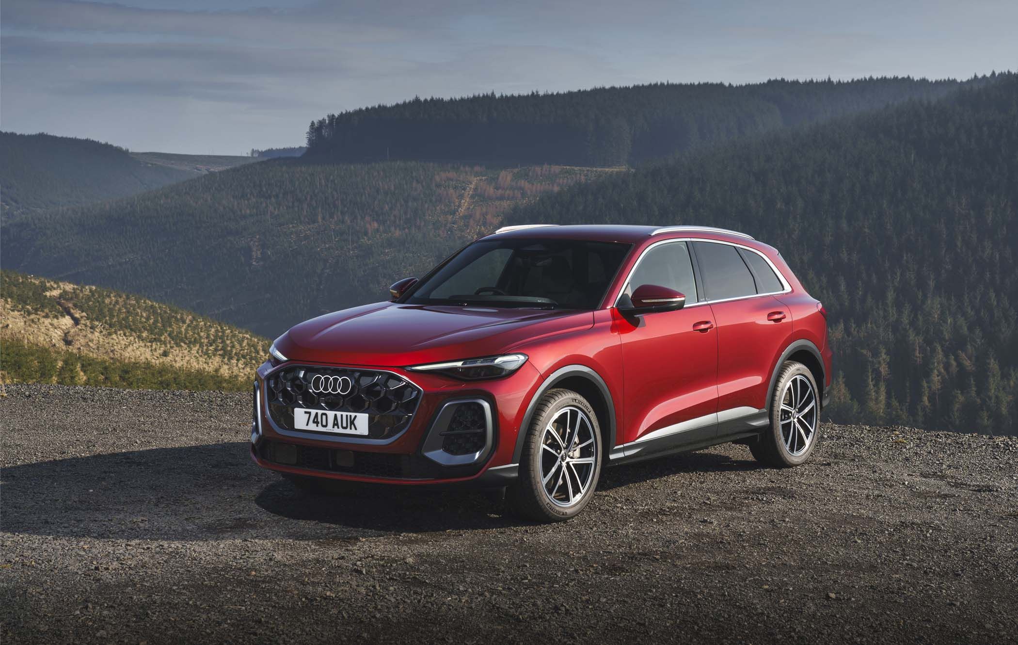 Red Audi Q5 parked on a gravel road, with a scenic backdrop of forested hills under a clear sky.