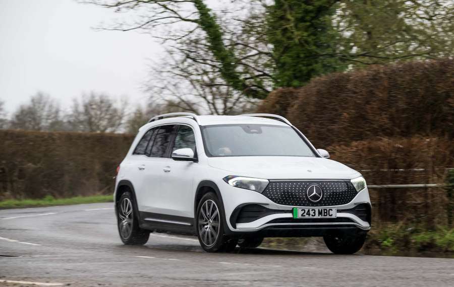 White Mercedes EQB driving on a wet, curvy road surrounded by leafless trees and hedges on a cloudy day.