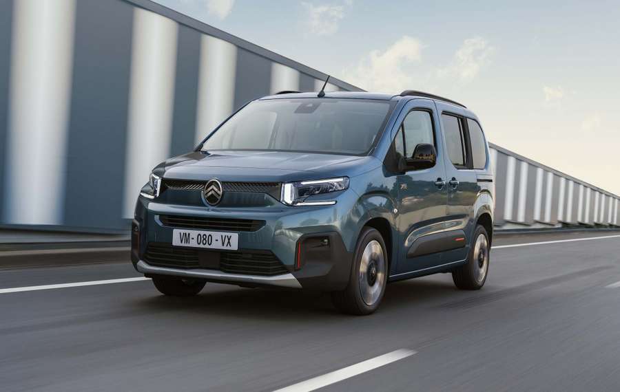 A blue Citroën ë-Berlingo driving on a highway with a modern bridge in the background under a clear sky.