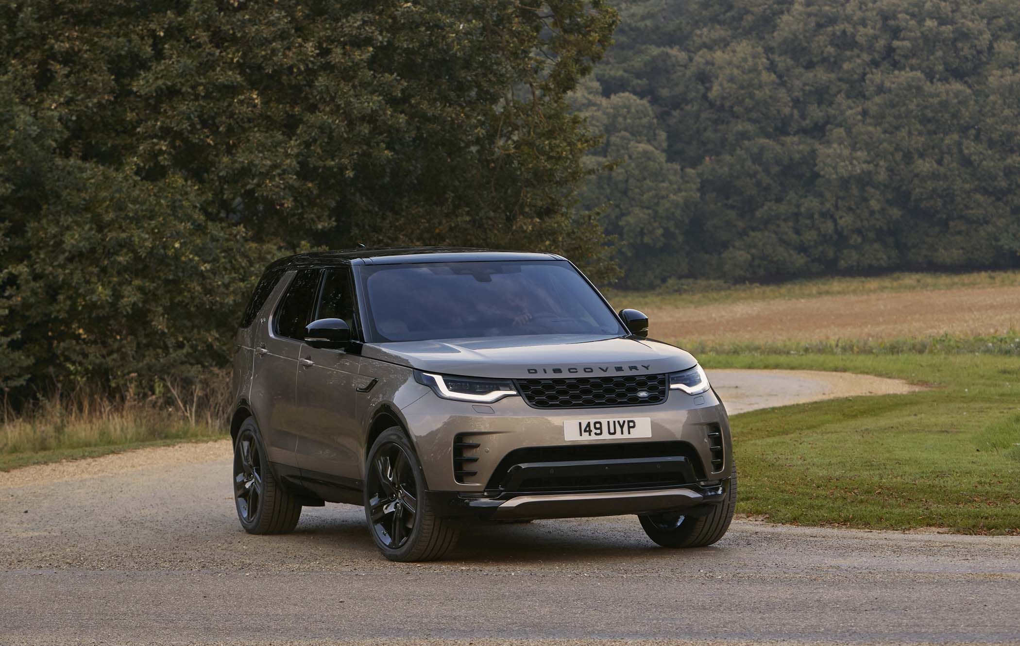Silver Land Rover Discovery parked on a gravel path beside lush greenery and trees, with a winding road in the background.