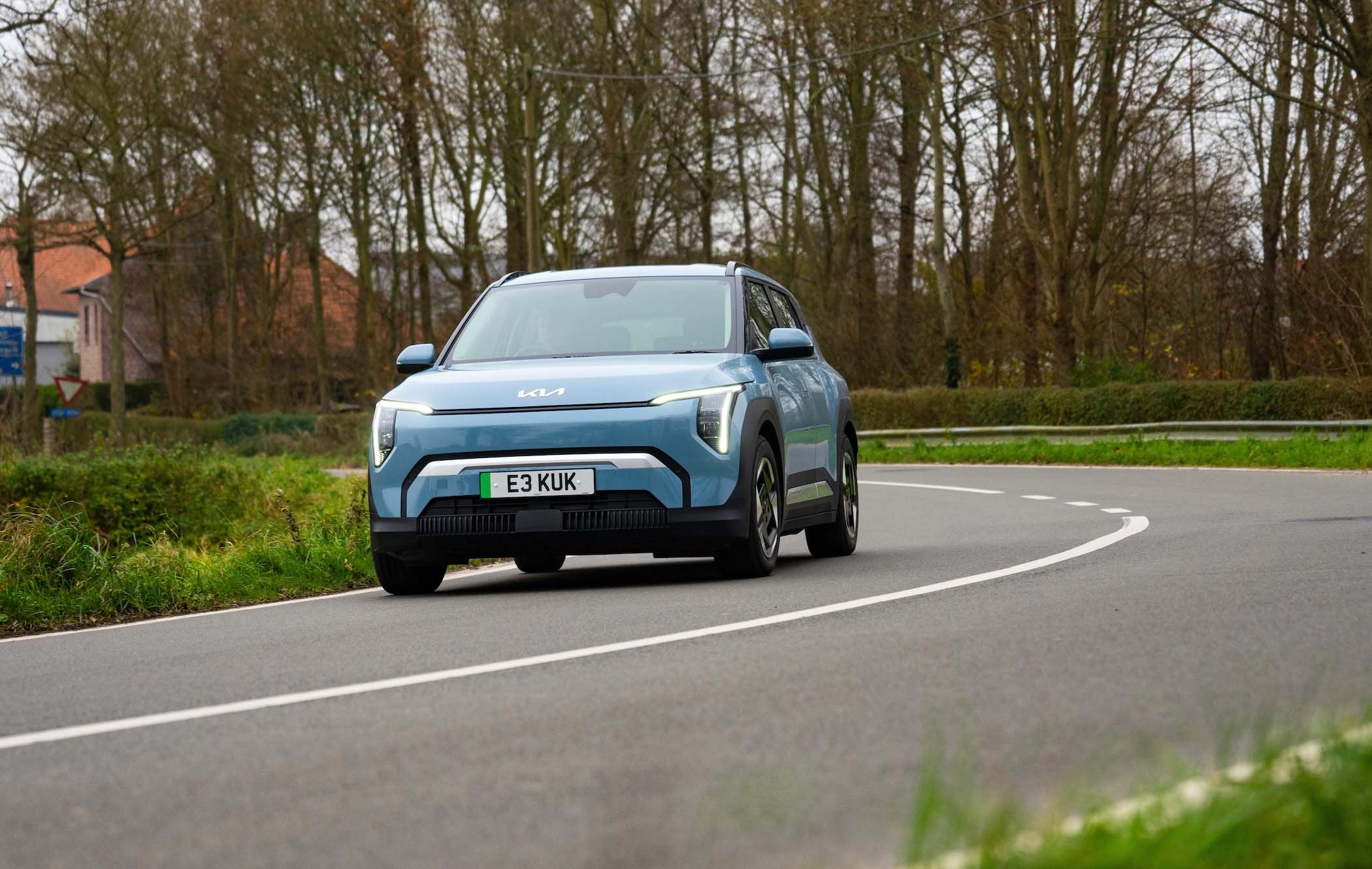 Blue Kia EV3 driving on a curved rural road, surrounded by trees and grass, with houses in the background.