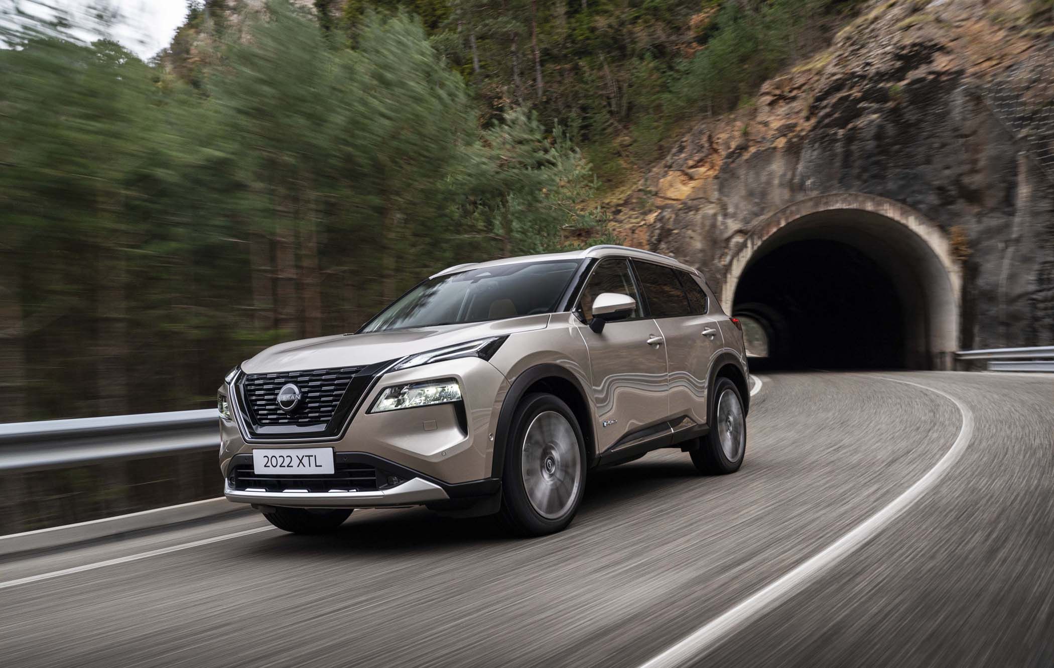 Nissan X-Trail driving through a mountainous road with a tunnel exit, surrounded by trees. The vehicle is glossy beige.