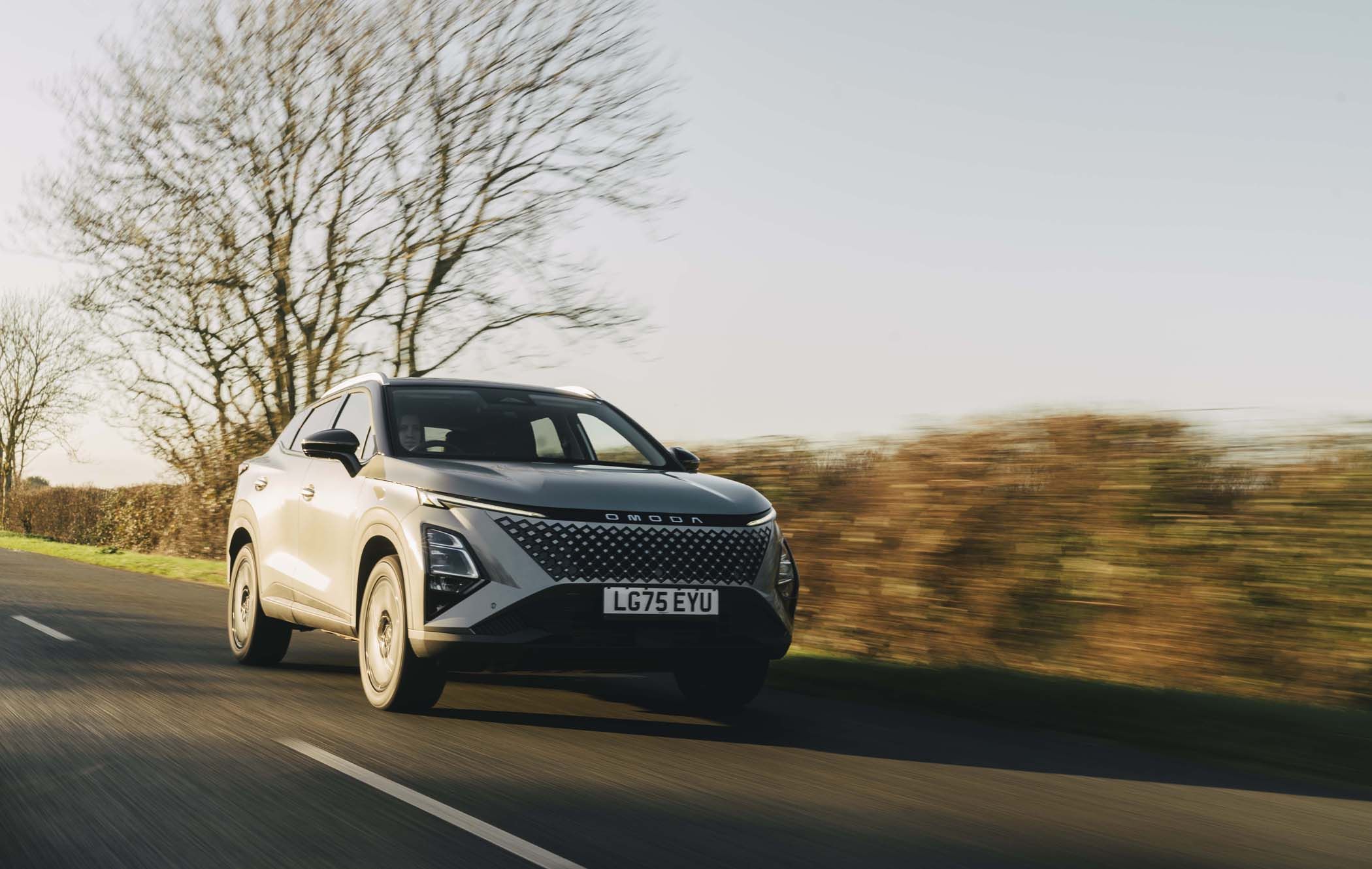 Silver OMODA 5 SHS-H driving on a rural road with blurred trees and hedges in the background, under a clear sky.