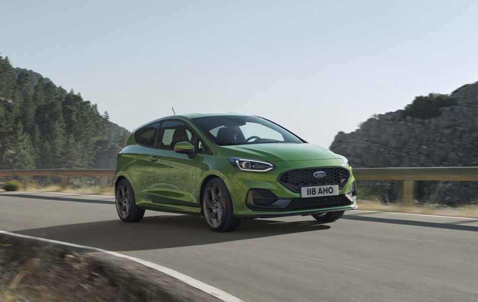 Green Ford Fiesta driving on a scenic mountain road with trees and rocky hills in the background under a clear sky.