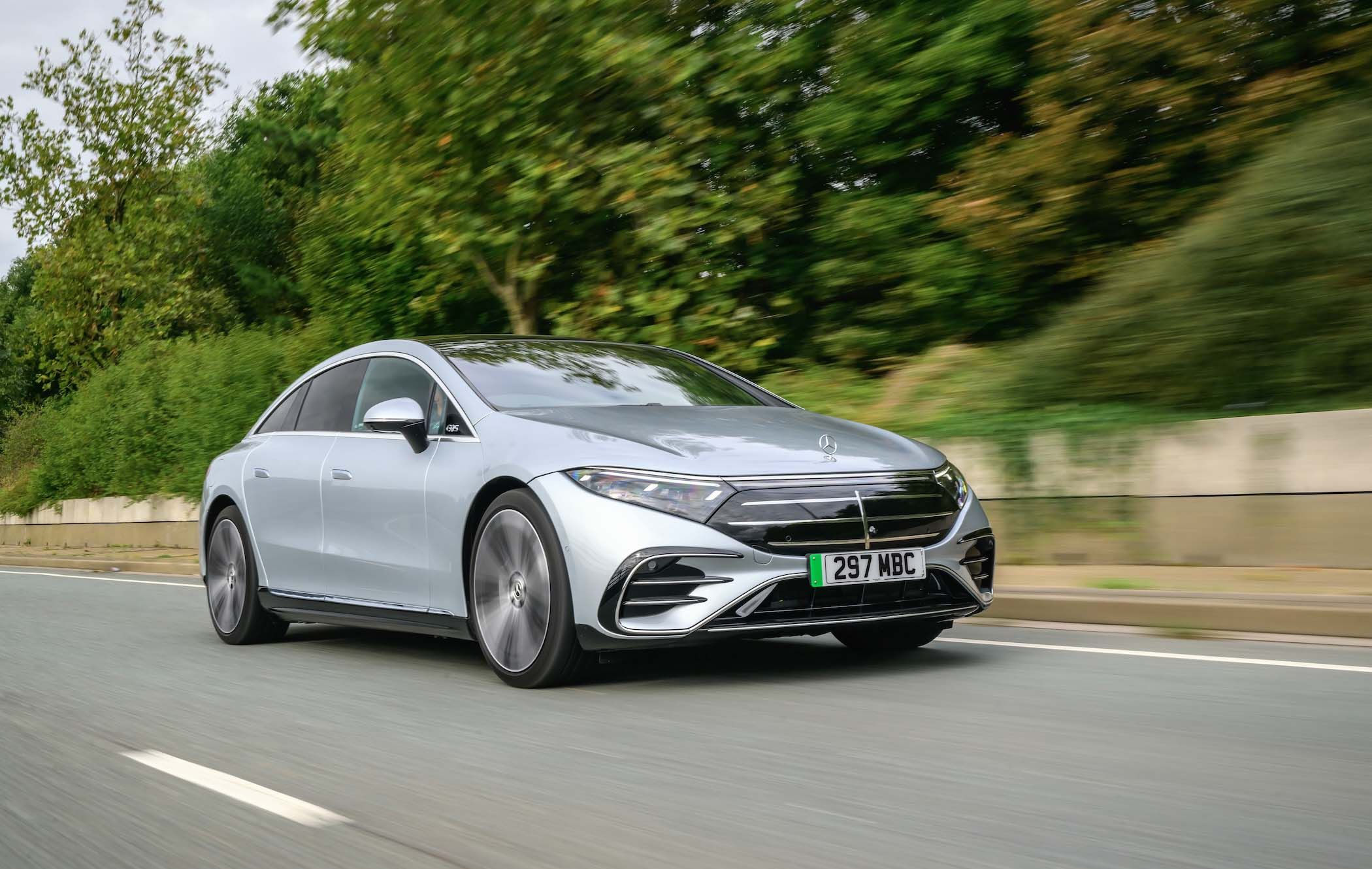 Silver Mercedes EQS driving on a road with blurred green foliage in the background, showcasing motion and speed.
