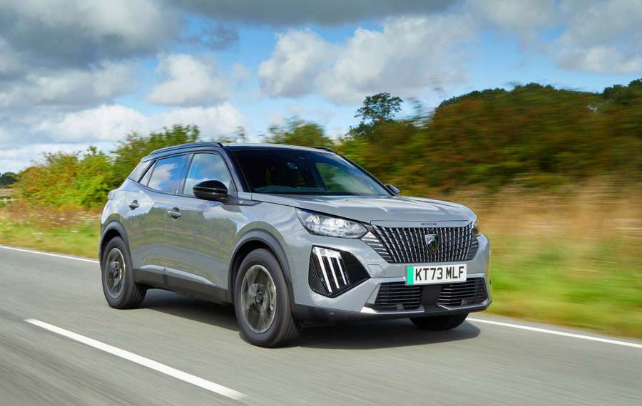 Silver Peugeot E-2008 driving on a countryside road with blurred greenery and cloudy sky in the background. License plate reads "KT73 MLF."