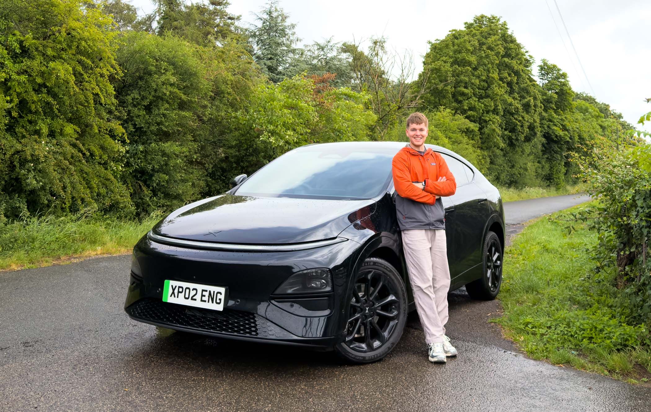 Craig Hale stands beside a black XPENG G6 on a wet road surrounded by lush green trees, wearing an orange jacket and smiling.