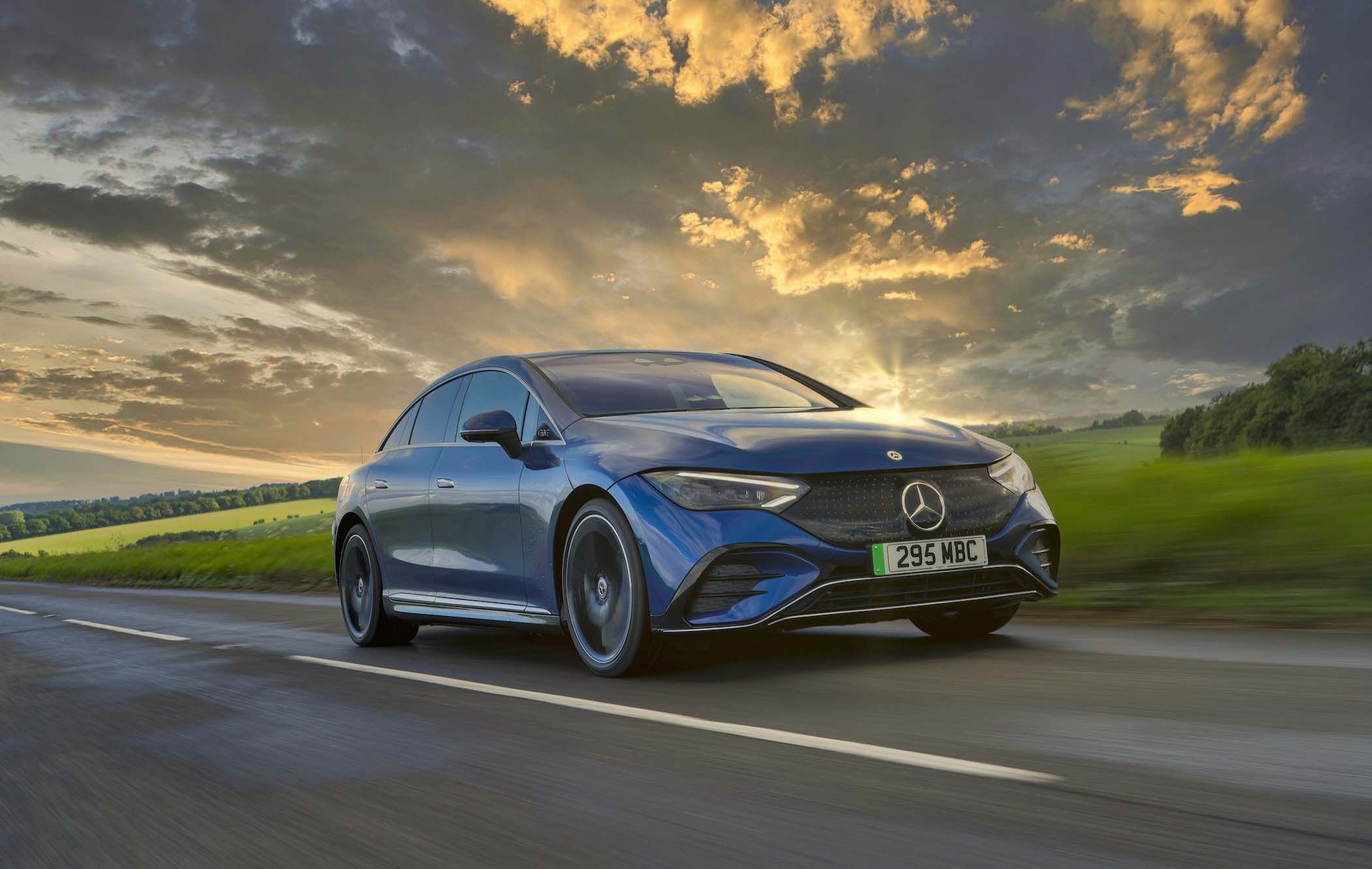 A blue Mercedes EQE driving on a countryside road at sunset, with dramatic clouds and green fields in the background.