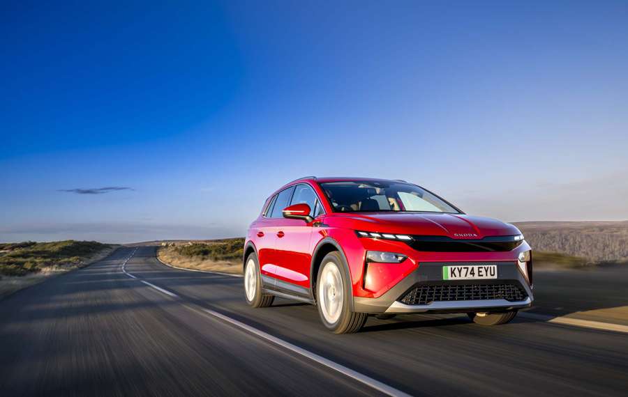 A red Škoda Elroq driving on a scenic, winding road under a clear blue sky, with blurred motion suggesting speed.