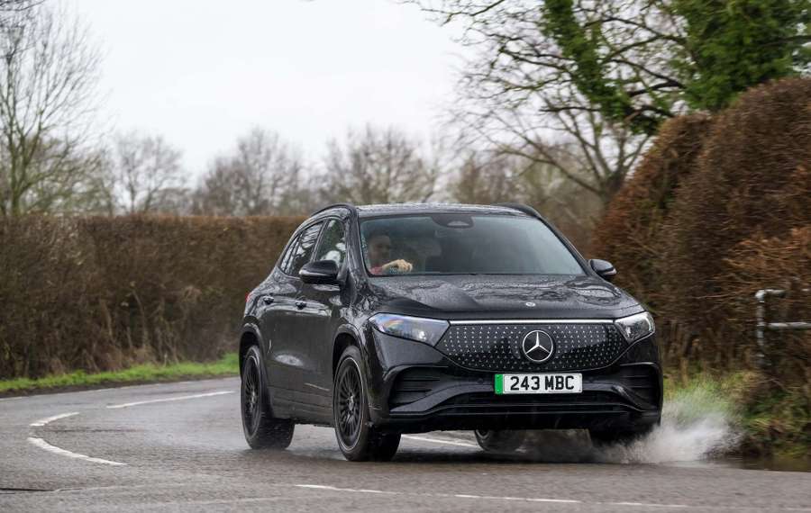 Black Mercedes EQA driving on a wet, curvy road, splashing water. Overcast sky and bare trees in the background.