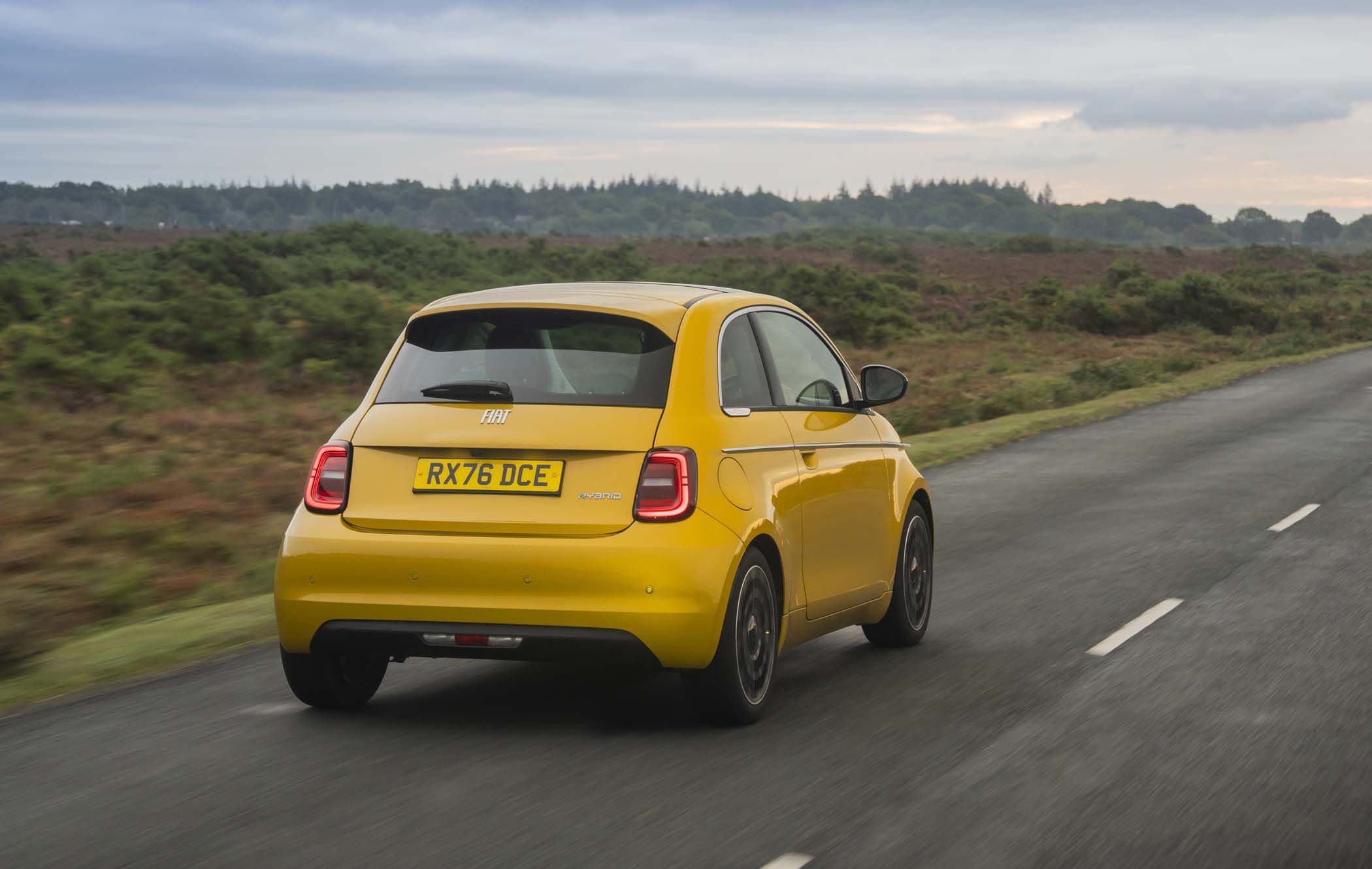 A yellow Fiat 500 Hybrid driving on a rural road with greenery and a cloudy sky in the background.