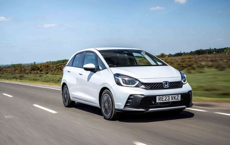 A white Honda Jazz driving on a rural road with greenery and a blue sky in the background.