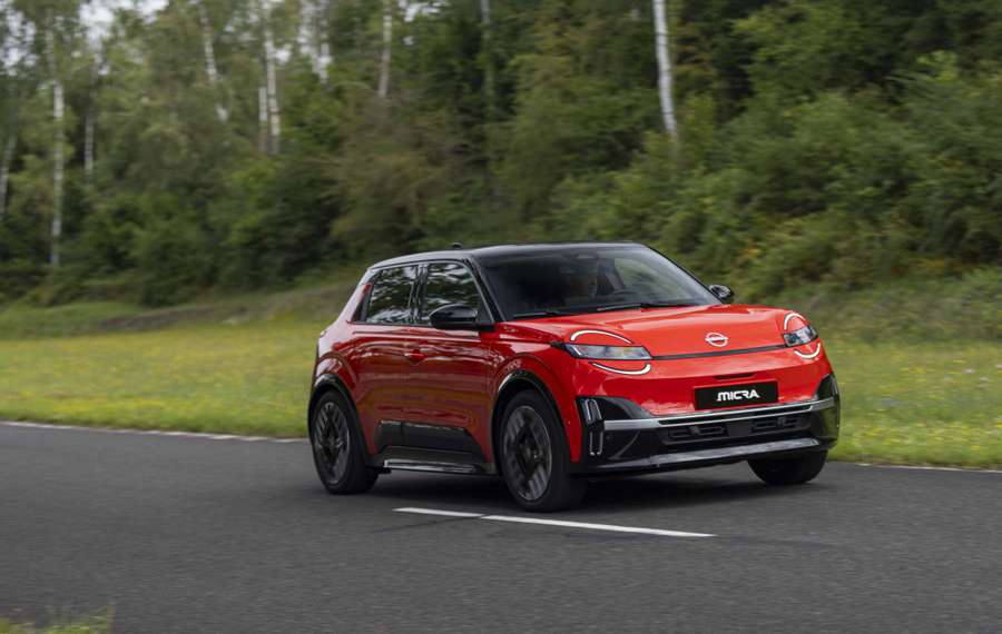 Red Nissan Micra driving on a road through a green, wooded area on a clear day.