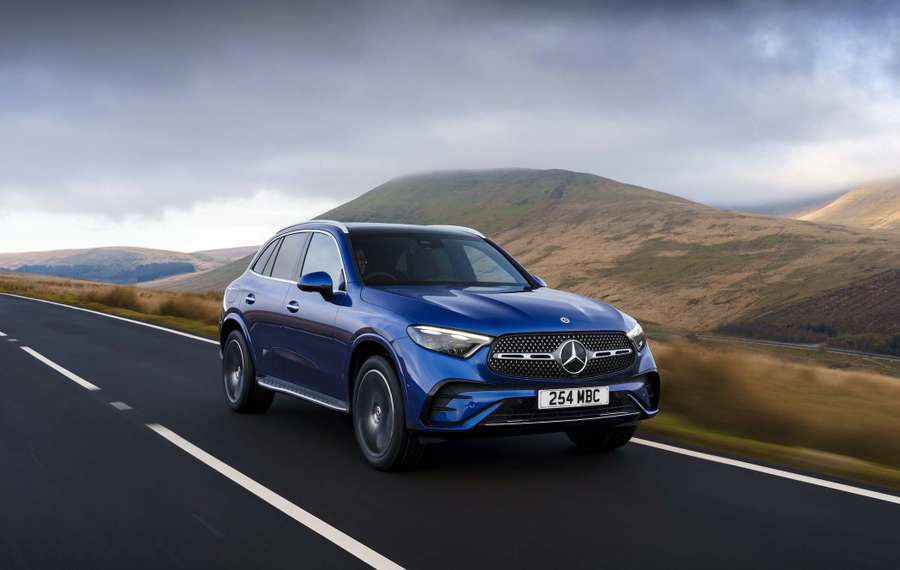 A blue Mercedes GLC drives on a scenic road with rolling hills and a cloudy sky in the background.