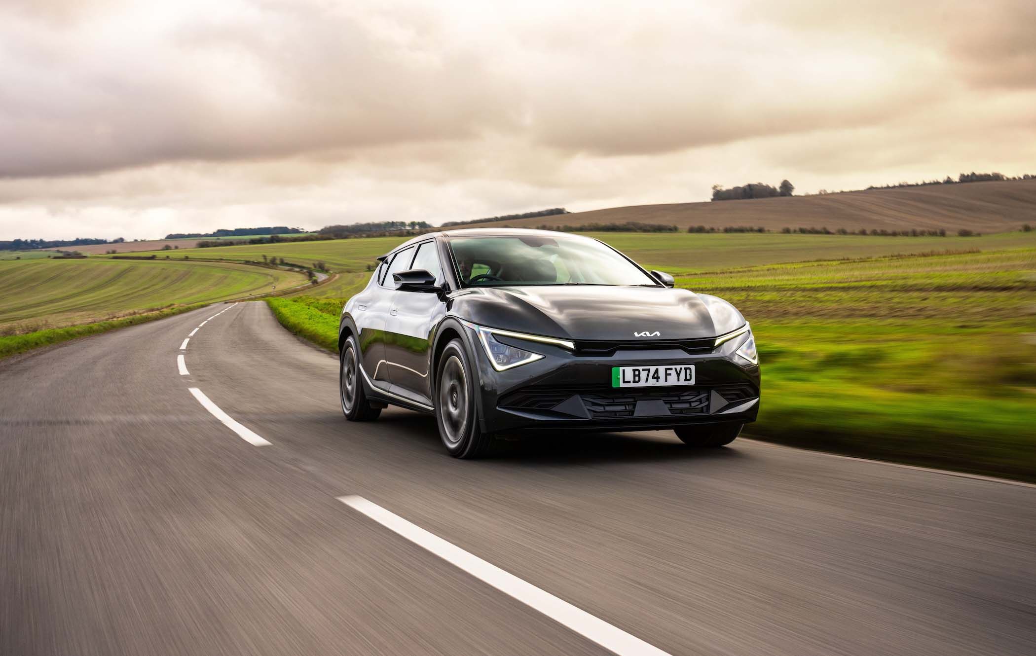 A black Kia EV6 drives on a winding rural road with green fields and a cloudy sky in the background.